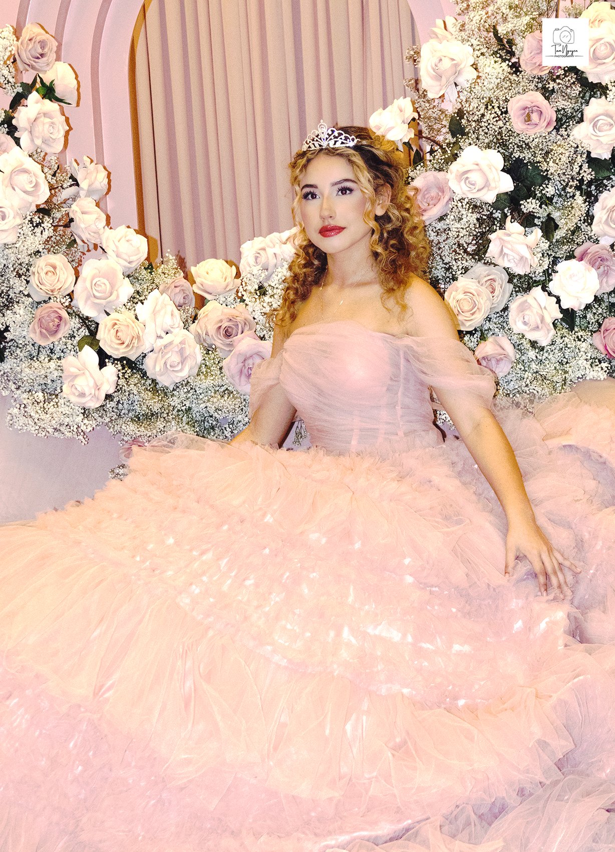 Plate six: a young woman in a pink quinceañera gown and tiara seated in front of a wall of roses and baby's breath.